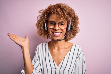African american curly call center agent woman working using headset over pink background smiling cheerful presenting and pointing with palm of hand looking at the camera.