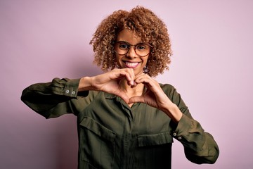Young beautiful african american woman wearing casual shirt and glasses over pink background smiling in love showing heart symbol and shape with hands. Romantic concept.