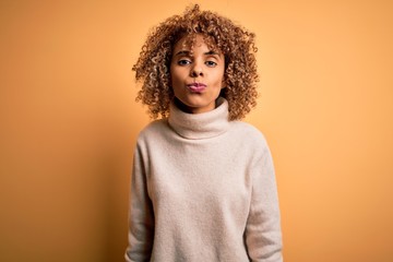 Young beautiful african american woman wearing turtleneck sweater over yellow background looking at the camera blowing a kiss on air being lovely and sexy. Love expression.