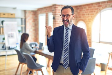Middle age handsome businessman wearing glasses   standing at the office smiling with happy face looking and pointing to the side with thumb up.