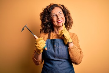 Middle age gardener woman wearing apron holding rake over isolated yellow background serious face thinking about question, very confused idea