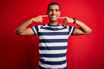 Handsome african american man wearing casual striped t-shirt standing over red background smiling cheerful showing and pointing with fingers teeth and mouth. Dental health concept.