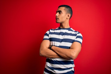 Handsome african american man wearing casual striped t-shirt standing over red background looking to the side with arms crossed convinced and confident