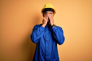 Young handsome african american worker man wearing blue uniform and security helmet rubbing eyes...