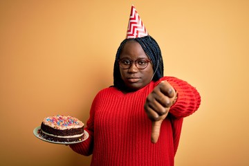 African american plus size woman wearinng birthday hat holding cake over yellow background with...