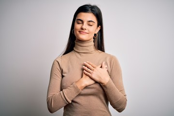 Young beautiful brunette woman wearing turtleneck sweater over white background smiling with hands on chest with closed eyes and grateful gesture on face. Health concept.