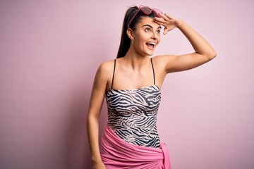 Young beautiful brunette woman on vacation wearing swimsuit over pink background very happy and smiling looking far away with hand over head. Searching concept.