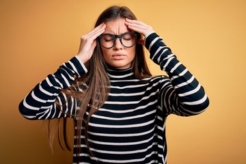Beautiful woman with blue eyes wearing striped sweater and glasses over yellow background suffering from headache desperate and stressed because pain and migraine. Hands on head.