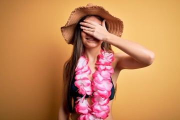 Young beautiful woman with blue eyes on vacation wearing bikini and hawaiian lei smiling and laughing with hand on face covering eyes for surprise. Blind concept.