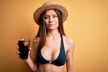 Young beautiful woman with blue eyes on vacation wearing bikini and hat drinking cola beverage with a confident expression on smart face thinking serious