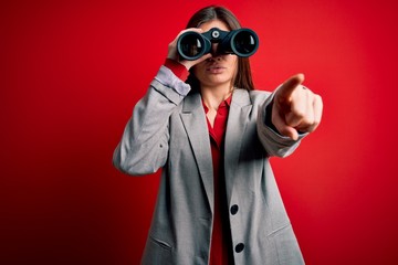 Young beautiful woman with blue eyes using binoculars over isolated red background pointing with...