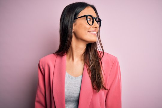 Young beautiful brunette businesswoman wearing jacket and glasses over pink background looking away to side with smile on face, natural expression. Laughing confident.