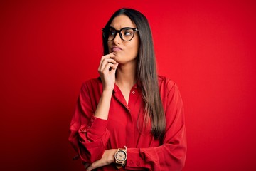 Young beautiful brunette woman wearing casual shirt and glasses over red background with hand on...