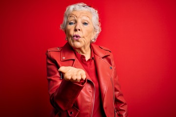 Senior beautiful grey-haired woman wearing casual red jacket and sunglasses looking at the camera blowing a kiss with hand on air being lovely and sexy. Love expression. - obrazy, fototapety, plakaty