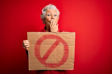 Senior beautiful grey-haired woman holding banner with prohibited signal over red background cover mouth with hand shocked with shame for mistake, expression of fear, scared in silence, secret concept