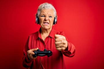 Senior beautiful grey-haired gamer woman playing video game using joystick and headphones annoyed and frustrated shouting with anger, crazy and yelling with raised hand, anger concept © Krakenimages.com