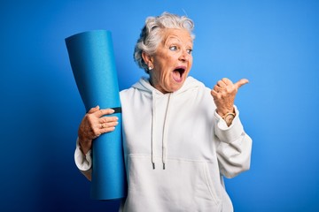 Senior beautiful sporty woman holding mat for yoga standing over isolated blue background pointing and showing with thumb up to the side with happy face smiling - obrazy, fototapety, plakaty
