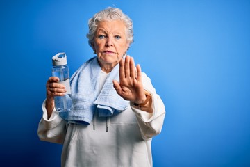 Senior beautiful sporty woman holding bottle of water standing over isolated blue background with open hand doing stop sign with serious and confident expression, defense gesture