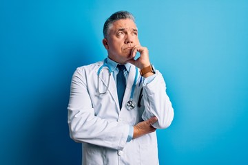 Middle age handsome grey-haired doctor man wearing coat and blue stethoscope with hand on chin...