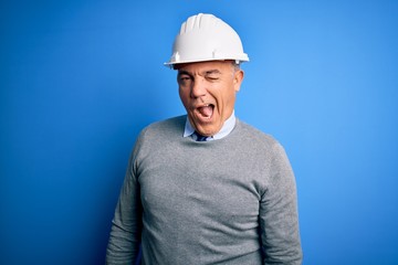 Middle age handsome grey-haired engineer man wearing safety helmet over blue background winking...