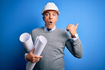 Middle age handsome grey-haired architect man wearing safety helmet holding blueprints Surprised pointing with hand finger to the side, open mouth amazed expression.