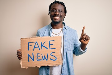 Young african american man holding banner with fake news message over white background surprised...
