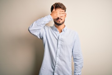 Young handsome man with beard wearing striped shirt standing over white background covering eyes with hand, looking serious and sad. Sightless, hiding and rejection concept