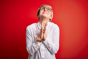 Young blonde business woman with short hair wearing glasses over red background begging and praying...