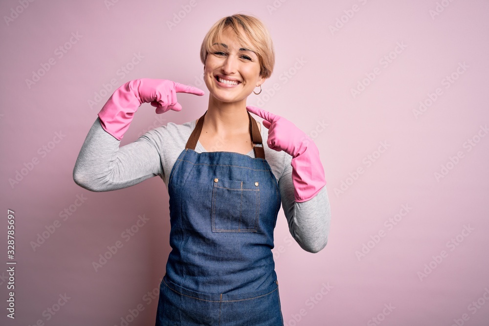 Poster young blonde cleaner woman with short hair wearing apron and gloves over pink background smiling che