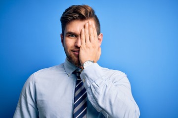 Young blond businessman with beard and blue eyes wearing elegant shirt and tie standing covering one eye with hand, confident smile on face and surprise emotion.