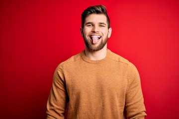 Young blond man with beard and blue eyes wearing casual s over red background sticking tongue out happy with funny expression. Emotion concept.