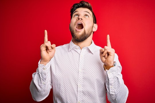 Young blond businessman with beard and blue eyes wearing elegant shirt over red background amazed and surprised looking up and pointing with fingers and raised arms.
