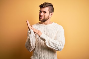 Young blond man with beard and blue eyes wearing white sweater over yellow background clapping and...