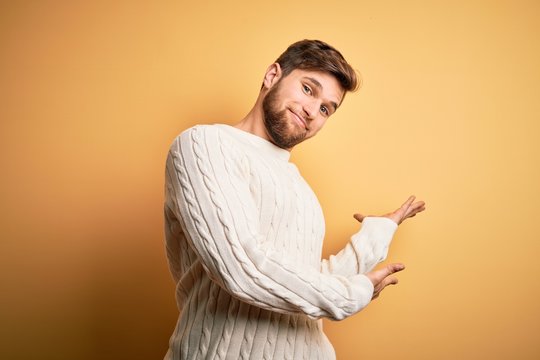 Young blond man with beard and blue eyes wearing white sweater over yellow background Inviting to enter smiling natural with open hand