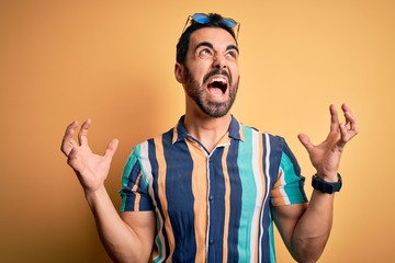 Handsome tourist man with beard on vacation wearing summer striped shirt and sunglasses crazy and mad shouting and yelling with aggressive expression and arms raised. Frustration concept.