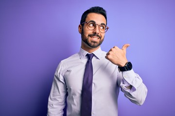 Handsome businessman with beard wearing casual tie and glasses over purple background smiling with happy face looking and pointing to the side with thumb up.