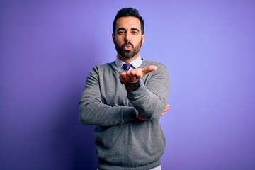 Handsome businessman with beard wearing casual tie standing over purple background looking at the camera blowing a kiss with hand on air being lovely and sexy. Love expression.
