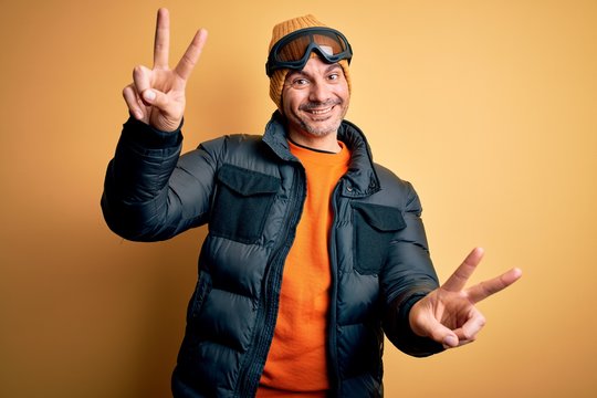 Young handsome skier man skiing wearing snow sportswear using ski goggles smiling with tongue out showing fingers of both hands doing victory sign. Number two.