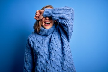Obraz premium Young beautiful blonde woman wearing casual turtleneck sweater over blue background covering eyes with arm smiling cheerful and funny. Blind concept.