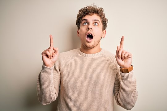 Young blond handsome man with curly hair wearing casual sweater over white background amazed and surprised looking up and pointing with fingers and raised arms.
