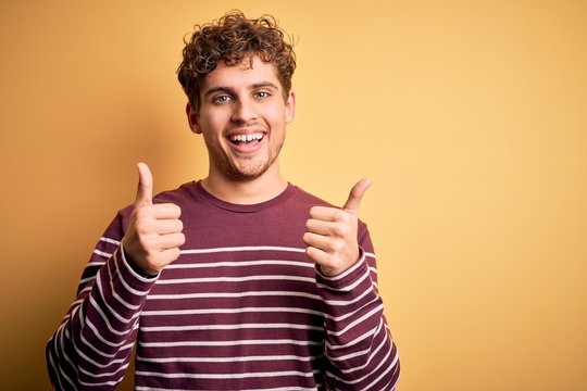 Young blond handsome man with curly hair wearing casual striped sweater success sign doing positive gesture with hand, thumbs up smiling and happy. Cheerful expression and winner gesture.