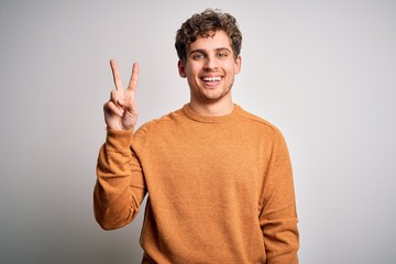 Young blond handsome man with curly hair wearing casual sweater over white background showing and pointing up with fingers number two while smiling confident and happy.