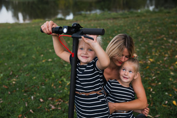 Twin blonde little girls sitting on electric scooter in park with their mother