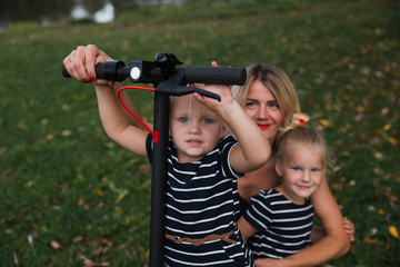Close up: Twin blonde little girls sitting on electric scooter in park with their mother