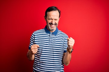 Middle age hoary man wearing casual striped polo standing over isolated red background excited for success with arms raised and eyes closed celebrating victory smiling. Winner concept.