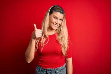 Obraz na płótnie Canvas Young beautiful blonde woman wearing casual t-shirt standing over isolated red background doing happy thumbs up gesture with hand. Approving expression looking at the camera showing success.