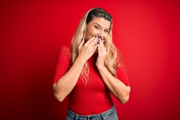 Young beautiful blonde woman wearing casual t-shirt standing over isolated red background laughing...