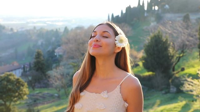 Relaxing and breathing fresh air in spring time. Beautiful young woman with eyes closed and flower on ear enjoying spring air in nature. Slow motion.