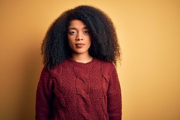 Young beautiful african american woman with afro hair standing over yellow isolated background with serious expression on face. Simple and natural looking at the camera.