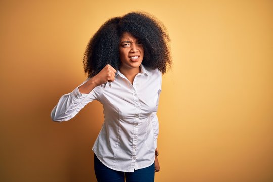 Young beautiful african american elegant woman with afro hair standing over yellow background angry and mad raising fist frustrated and furious while shouting with anger. Rage and aggressive concept.